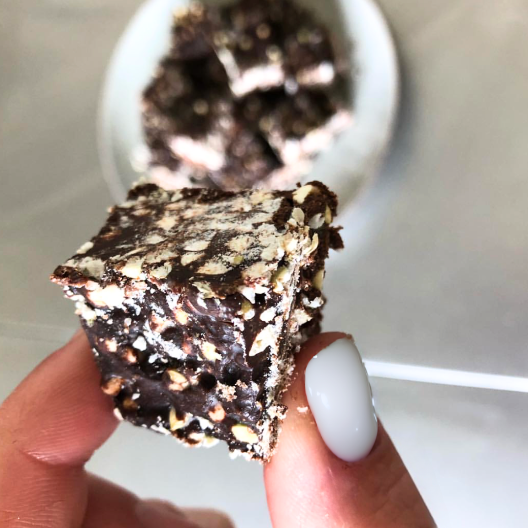 Close-up of Raw Lil Bucks fudge held between fingers showcasing puffed buckwheat clusters and glossy chocolate with a blurred bowl in the background