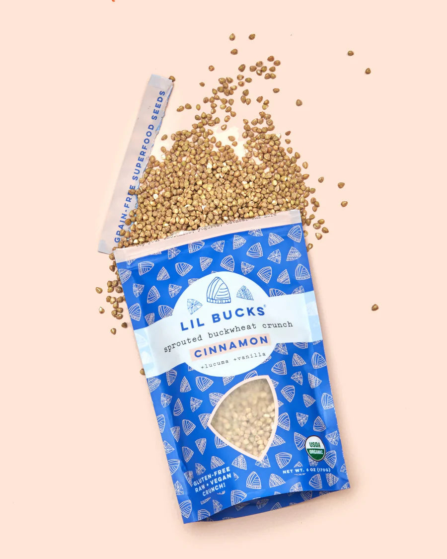Angled top view of Lil Bucks Cinnamon sprouted buckwheat crunch bag with spilled buckwheat on pale pink background