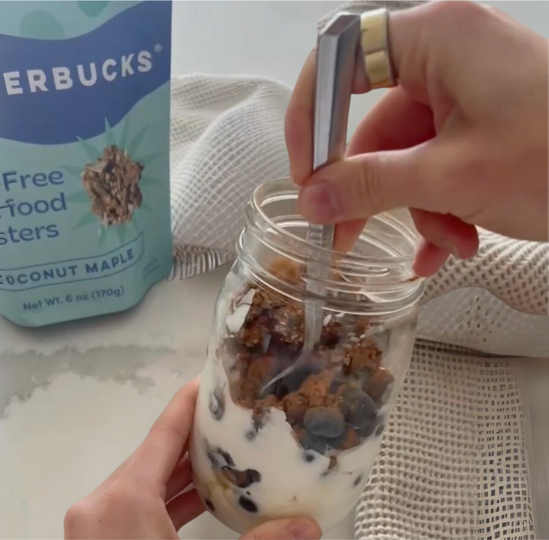 Hand stirring a mason jar of yogurt layered with blueberries and Coconut Maple Clusterbucks with product bag in background