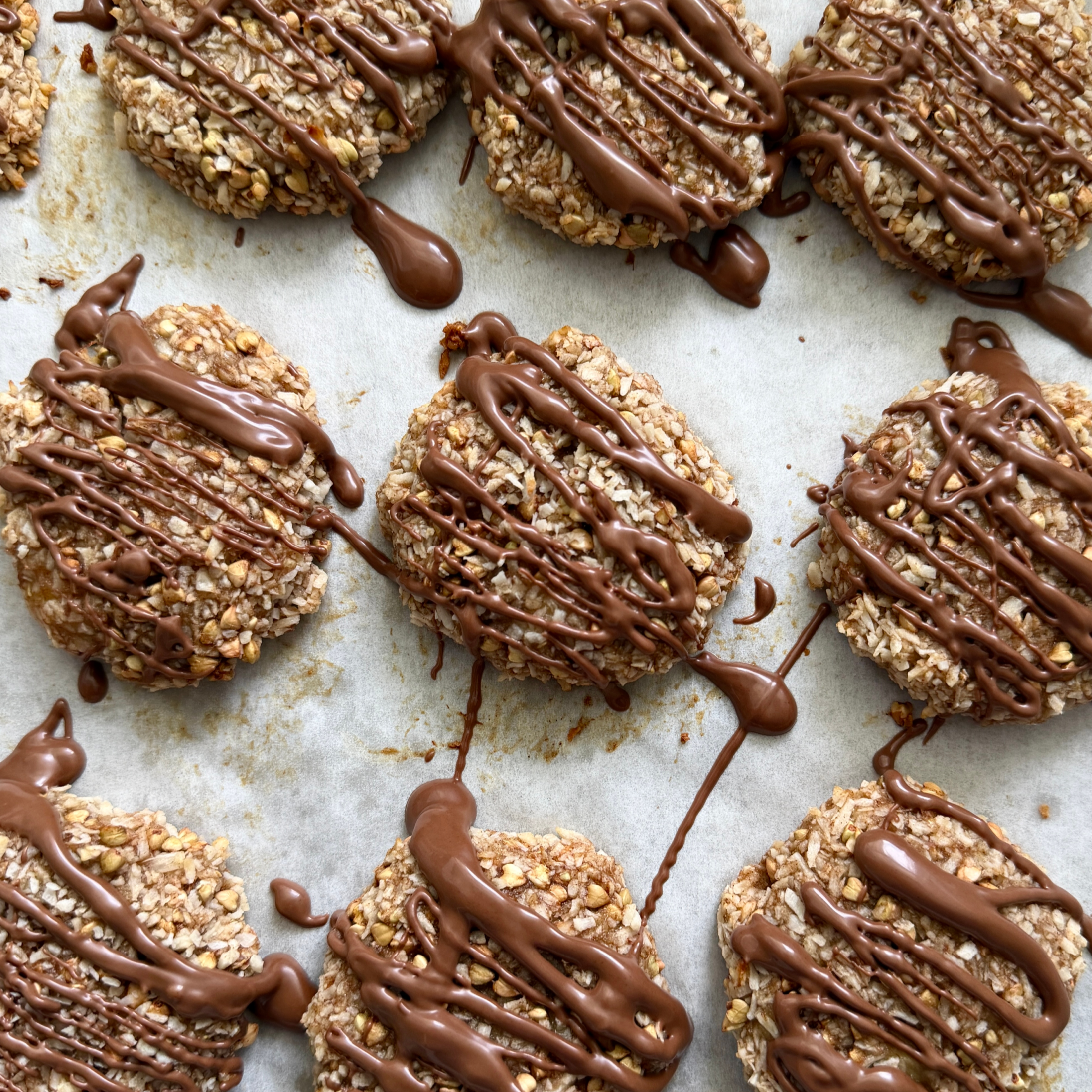 Top-down view of banana coconut gluten-free cookies drizzled with chocolate, showing oat flakes and toasted grains on parchment paper