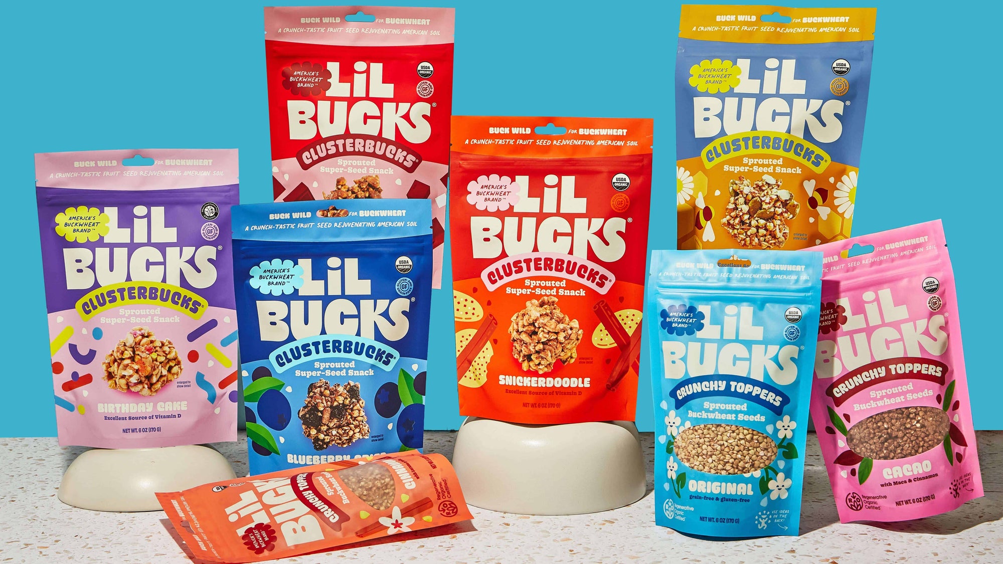 Assorted packages of Lil Bucks snack products on a light surface with a blue background